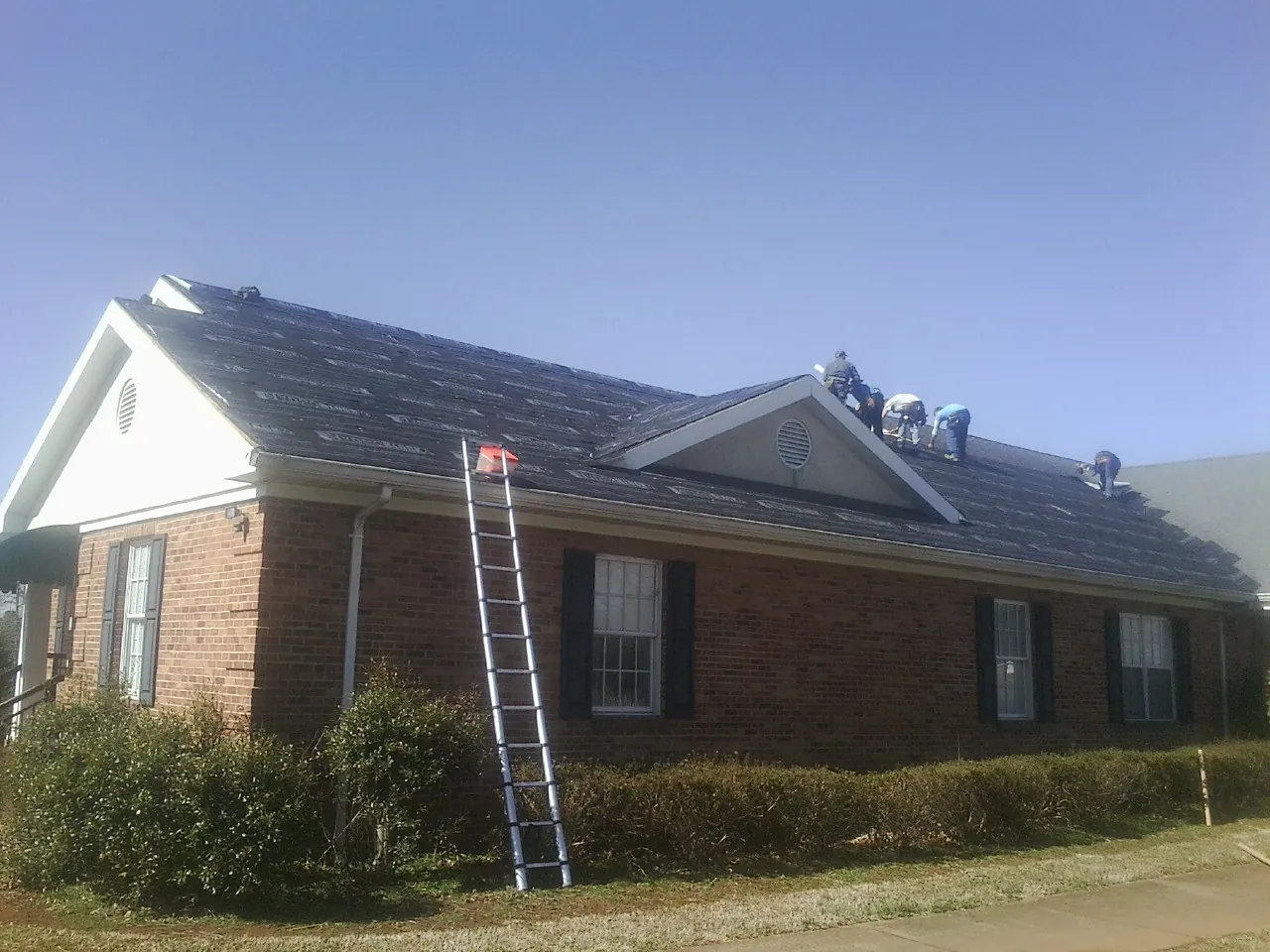 Crew tearing shingles off a brick ranch roof with a ladder set — Metal Roof Repair work by our crew in Nichols