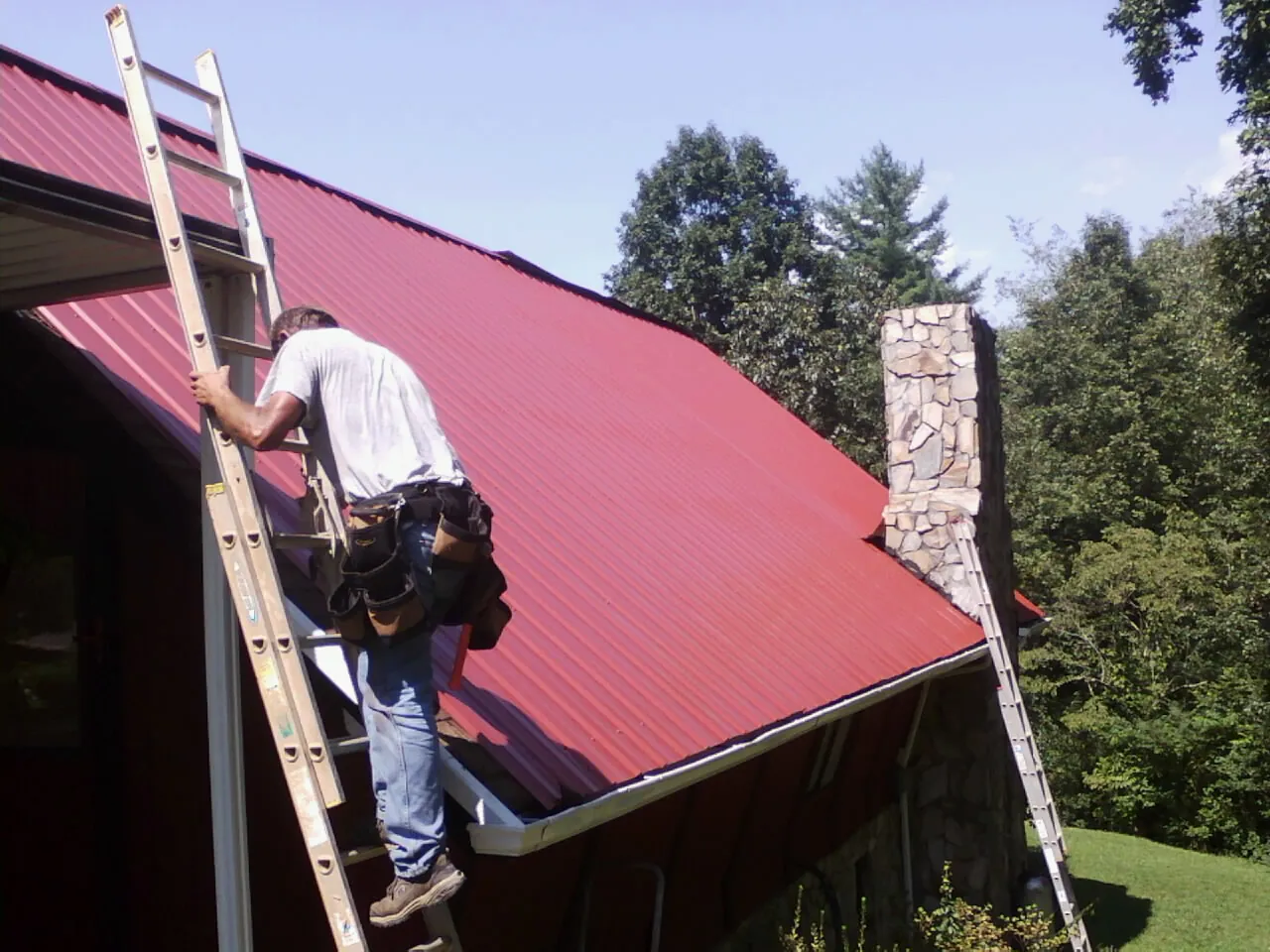 Roofer on a ladder beside a red metal roof and stone chimney — Soffit & Fascia Repair by Roofing Brothers in Nichols